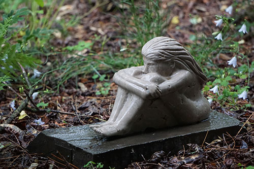 Image of a stone sculpture of a woman sitting in a contemplative stance among white flowers and rain.