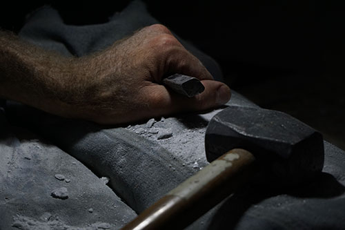 Image of a man's hand holding a chisel, next to a hammer and stone duct in low-light.